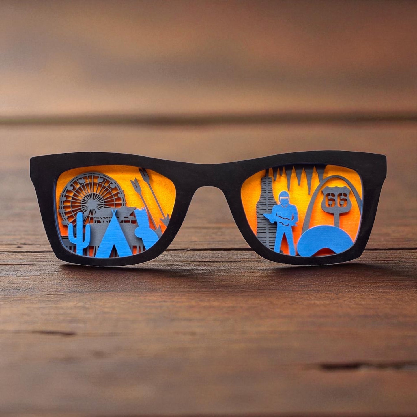 Black-framed wooden Route 66 skyline sunglasses wall decor standing upright on a wood surface, with layered orange and blue miniatures featuring the Santa Monica Pier, cactus, tepee, Cadillac Ranch, Twin Arrows, Pops on 66, Muffler Man, Missouri Caverns, the Cloud Gate, St. Louis Arch, and Route 66 sign.