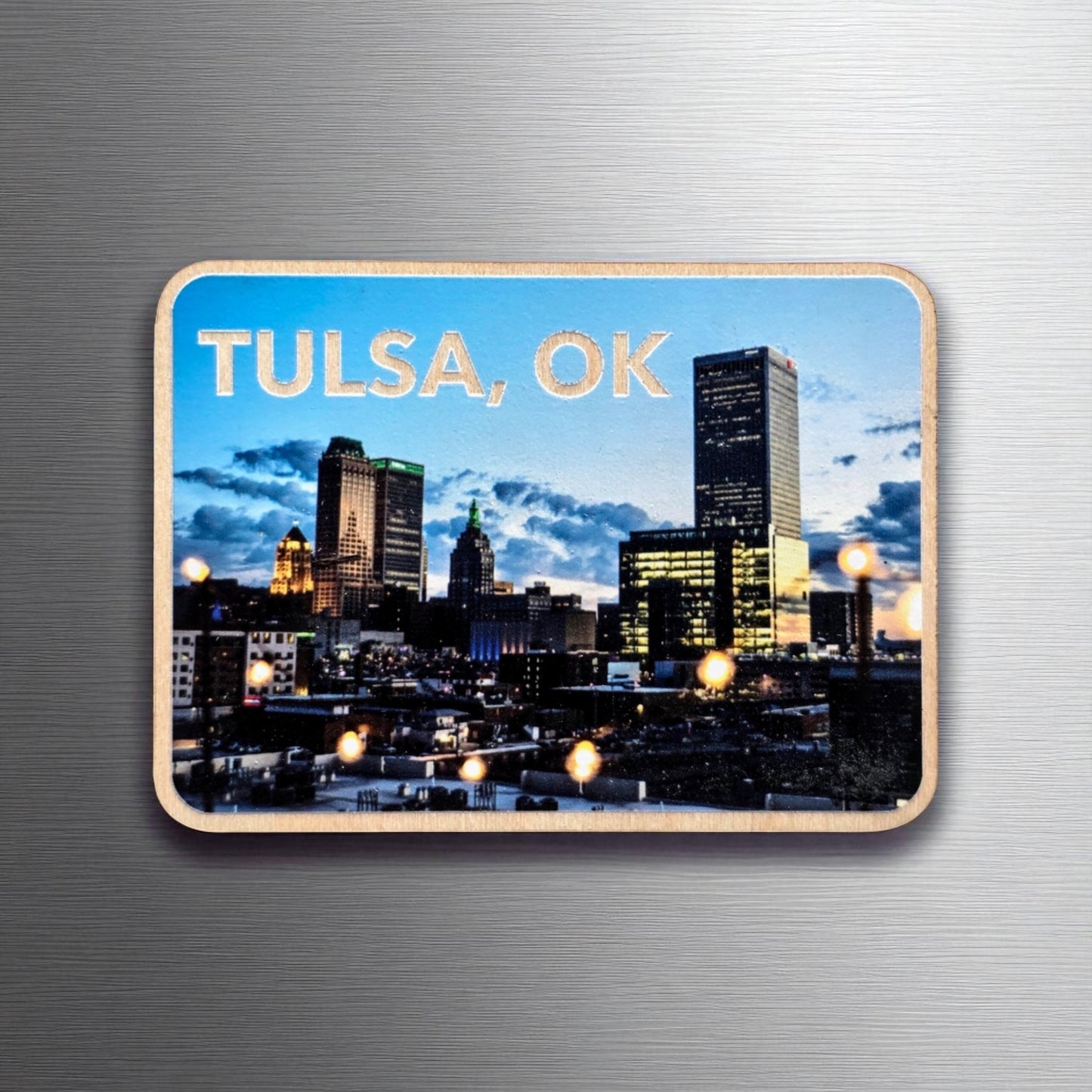 Wooden photo magnet featuring the downtown Tulsa, Oklahoma skyline at twilight. The buildings are lit up under a deep blue sky, with glowing string lights in the foreground. Displayed on a brushed metal surface.