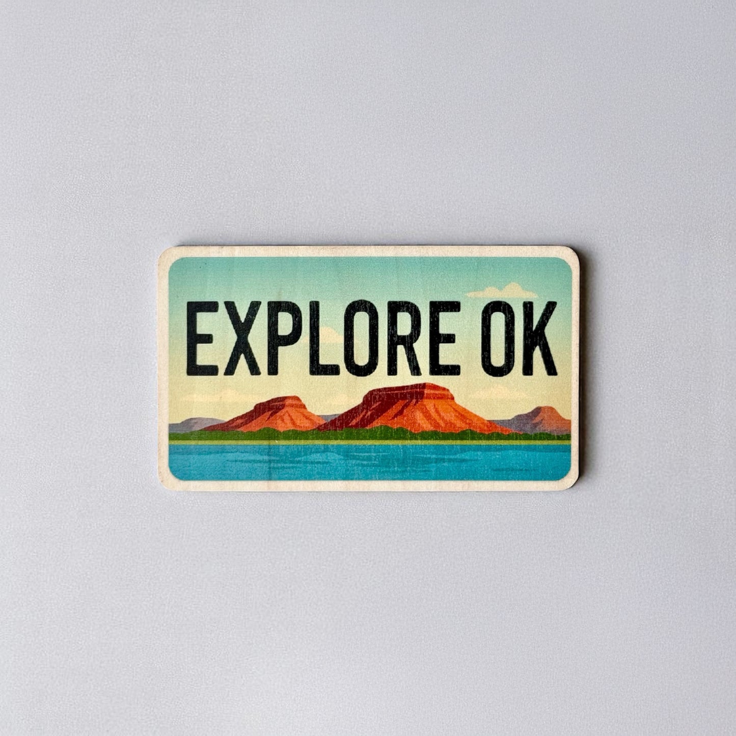 Vintage-style wood sticker that says “Explore OK” with red mesas and bright blue water, inspired by Oklahoma’s natural beauty.