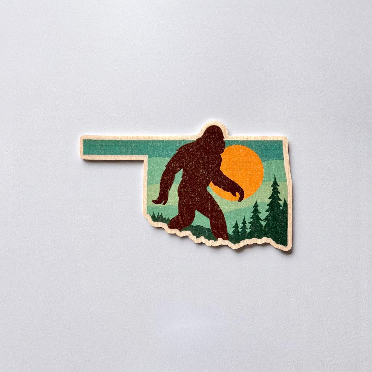 Oklahoma-shaped wood sticker featuring a silhouette of Bigfoot walking in front of a bold orange sunset with pine trees in the background β perfect for cryptid and Sasquatch fans.