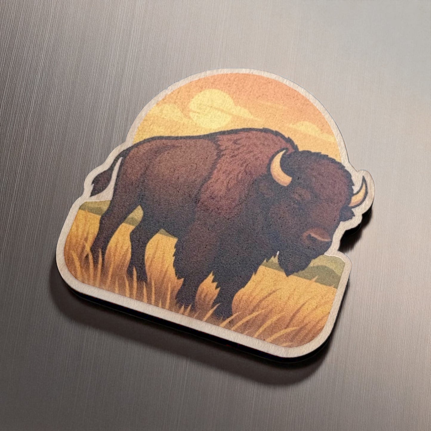 Angled view of maple bison magnet with bold silhouette and warm sunset printed on smooth wood surface against brushed metal