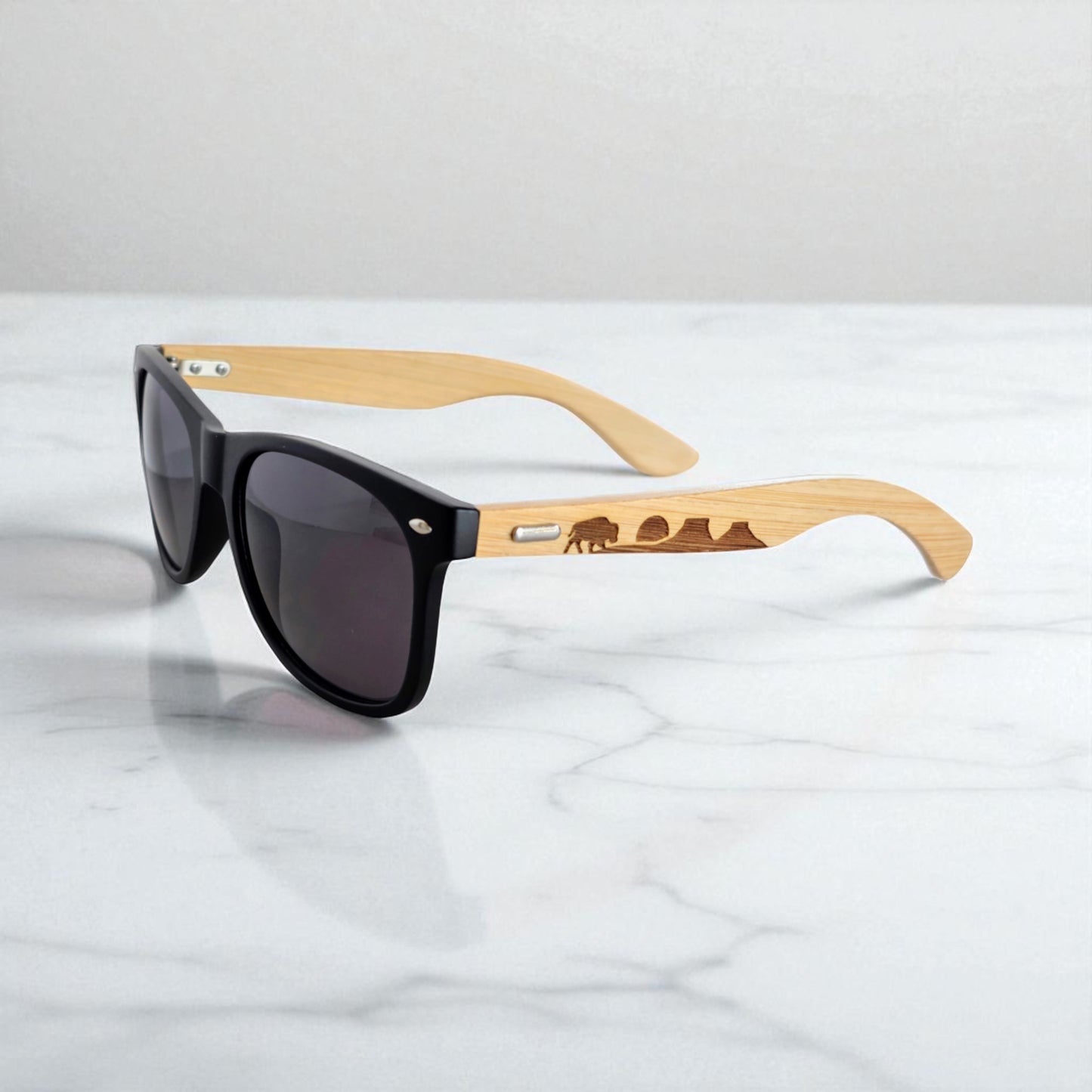 Oklahoma-themed bamboo sunglasses with a black frame and laser-engraved bison and mountain landscape, photographed on a marble background. A must-have accessory for nature lovers and proud Oklahomans wanting to showcase their love for wildlife, road trips, and rustic, sustainable style.
