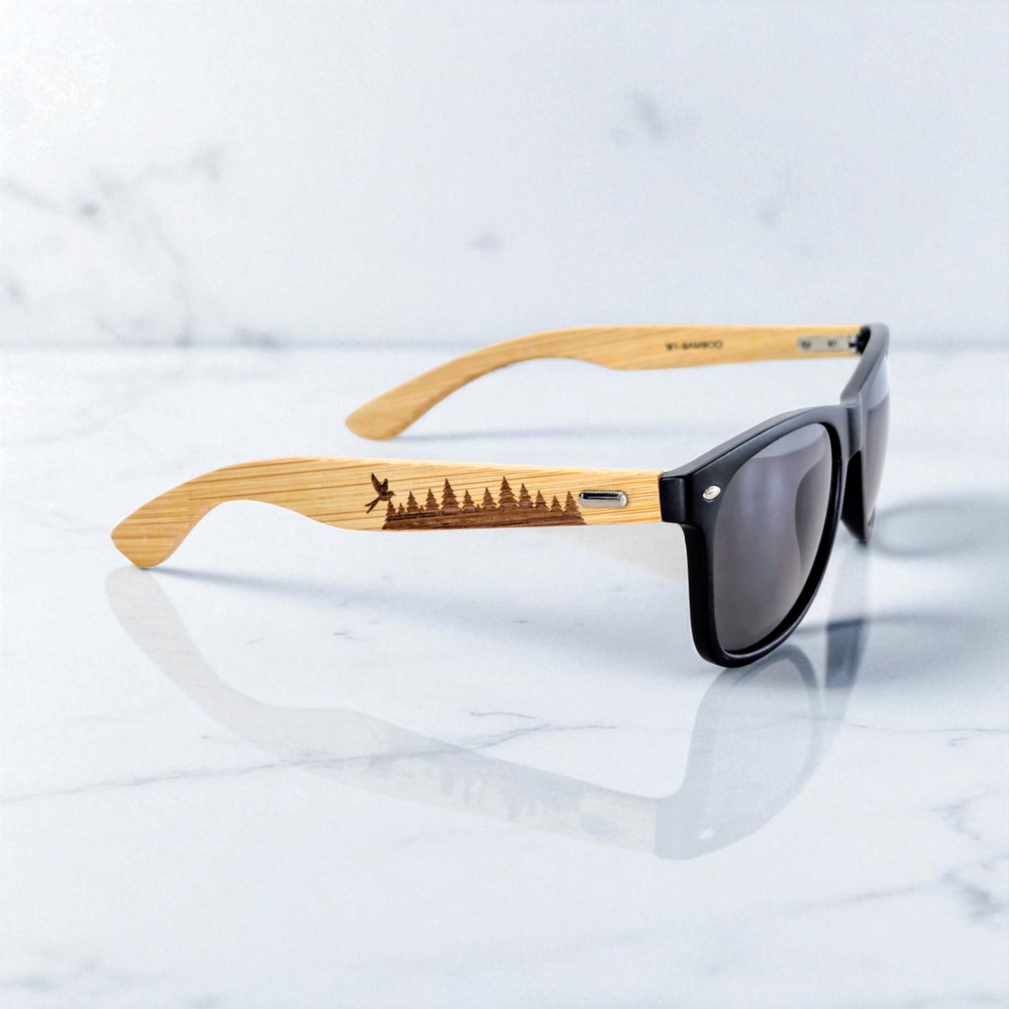 Adventure-ready sunglasses with bamboo arms engraved with an evergreen tree line and soaring bird, laid on a clean marble surface. This woodland-inspired design is perfect for Oklahoma hikers, lake-goers, and wildlife fans who want eyewear that blends function and forest vibes.
