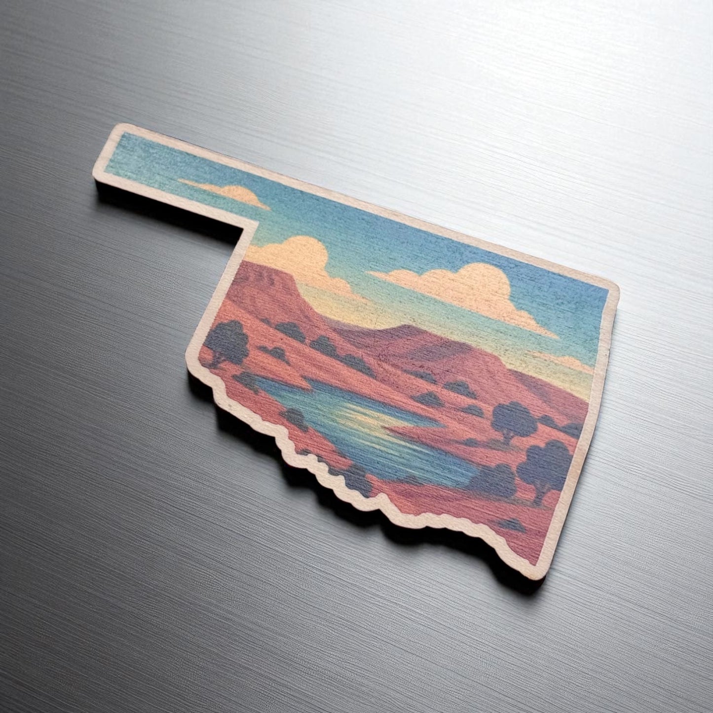 Angled view of Oklahoma shaped maple magnet with scenic red rock plateau design and vibrant sky on smooth brushed metal surface