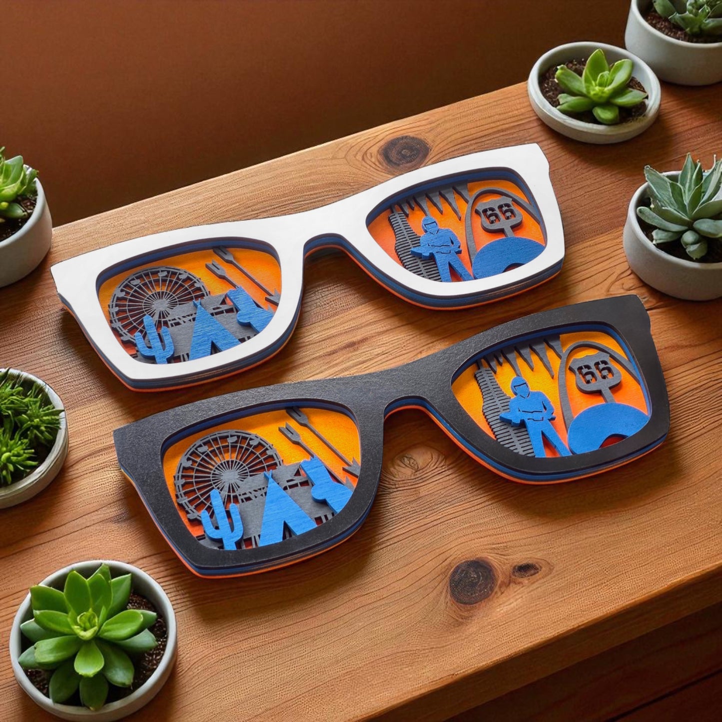 Flat lay of both white and black Route 66 wooden sunglasses on a wood surface, side by side, surrounded by small potted succulents, showing off the 3D layered skyline designs.