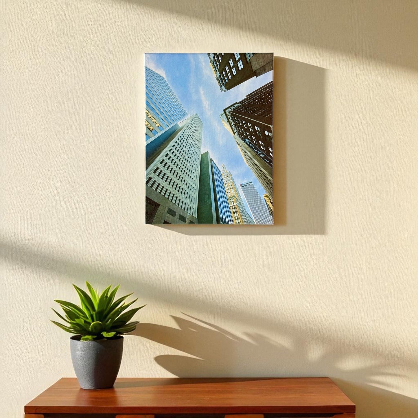 Vertical 12x16 inch Tulsa skyscraper canvas wall art shown hanging on a light-colored wall above a wooden table with a small green plant, bathed in afternoon light