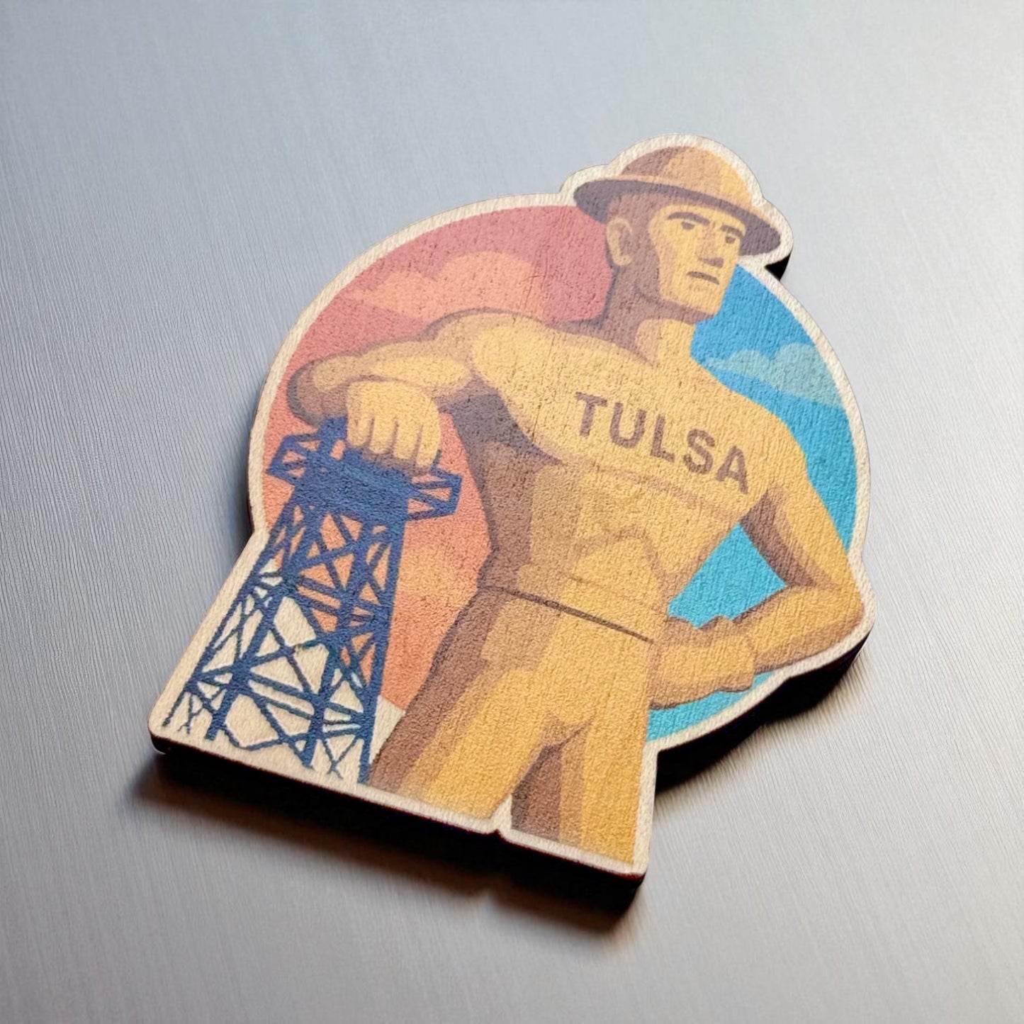 Angled view of Tulsa Golden Driller wood magnet showing cutout detail and vibrant sky design on brushed metal surface