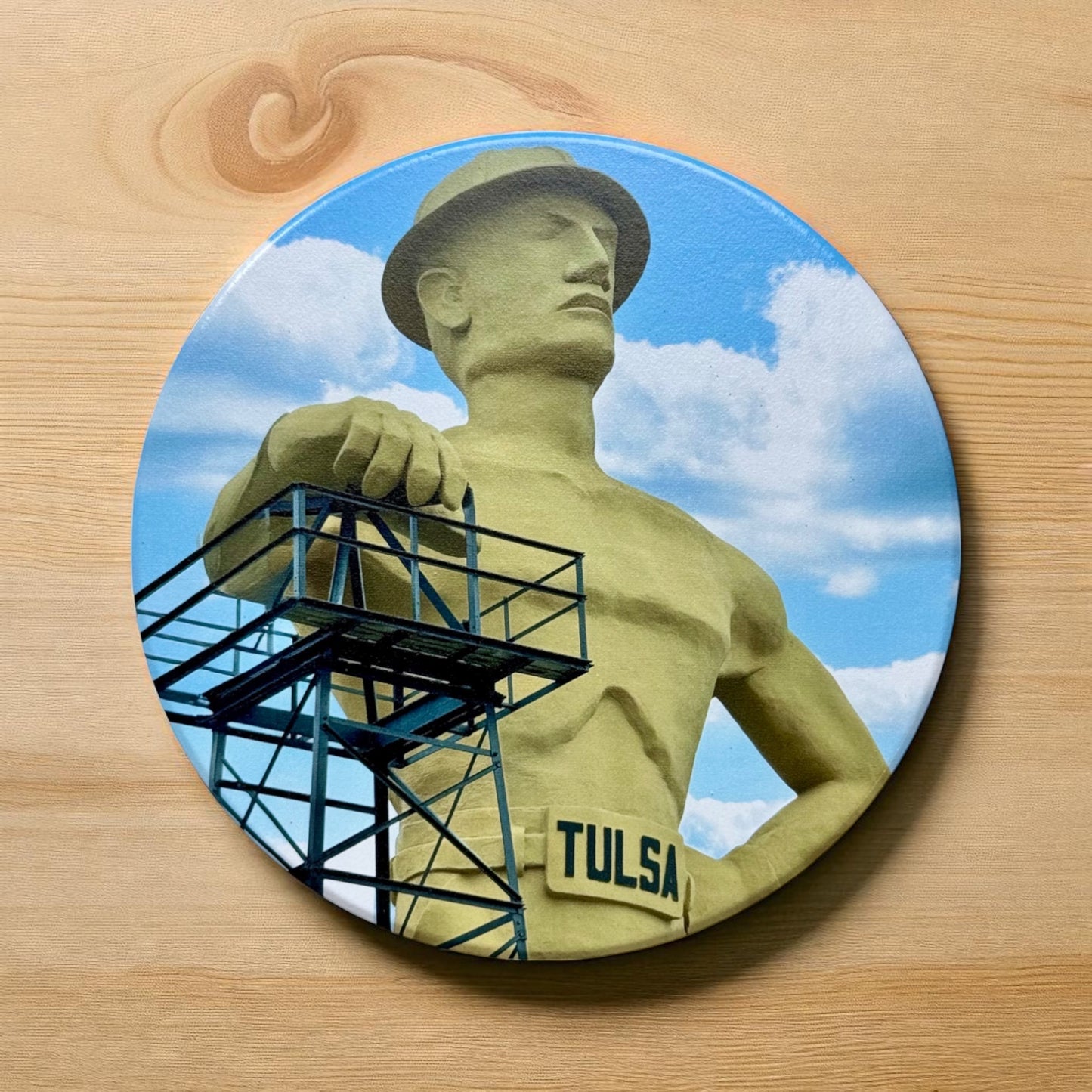 Colorful round ceramic coaster featuring Tulsa’s Golden Driller statue under a blue sky with white clouds, placed on a light wood surface