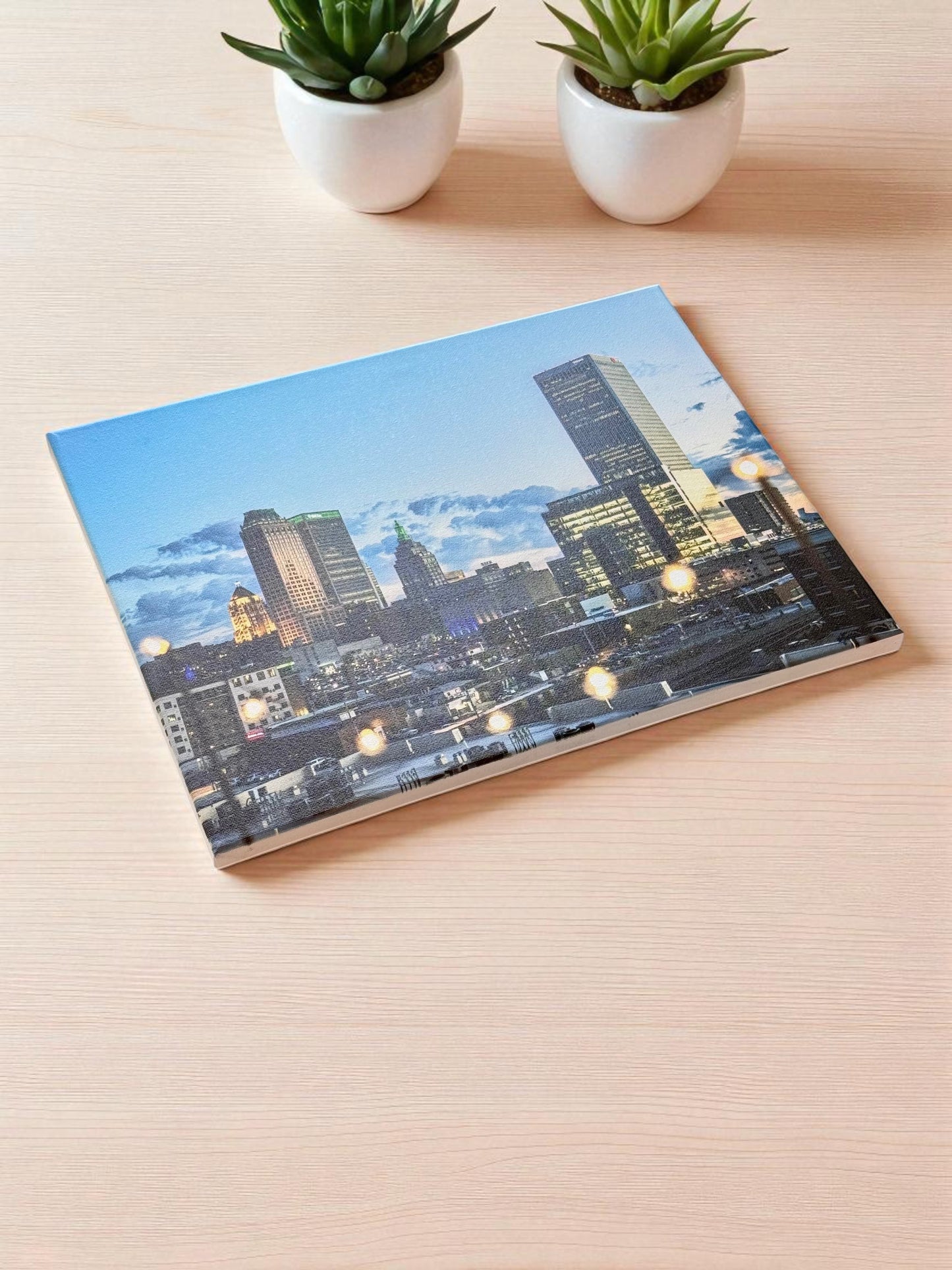 Flat-lay of a 12x16 inch Tulsa night skyline canvas on a light wooden surface, surrounded by two small potted succulents, showing vibrant evening colors and glowing city lights.