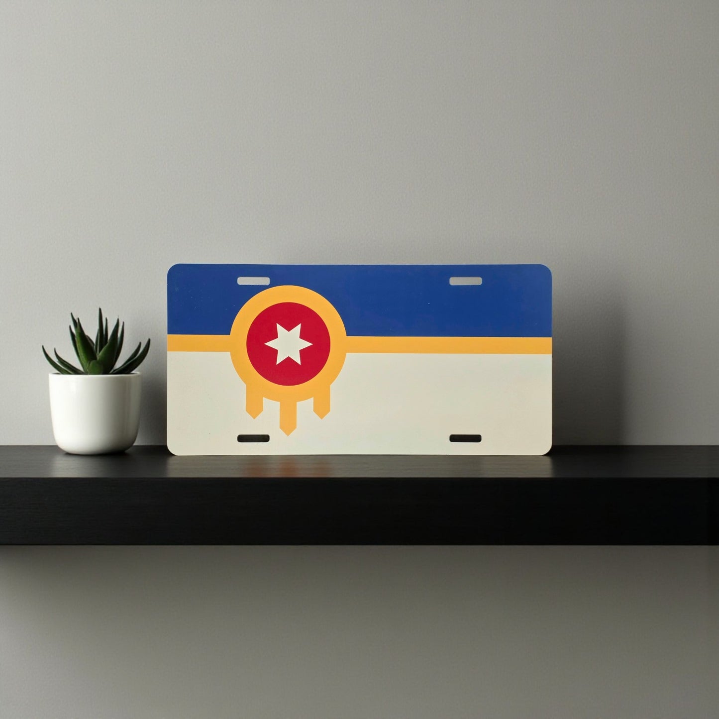 Tulsa flag vanity plate displayed on a black shelf against a gray wall with a small potted plant.