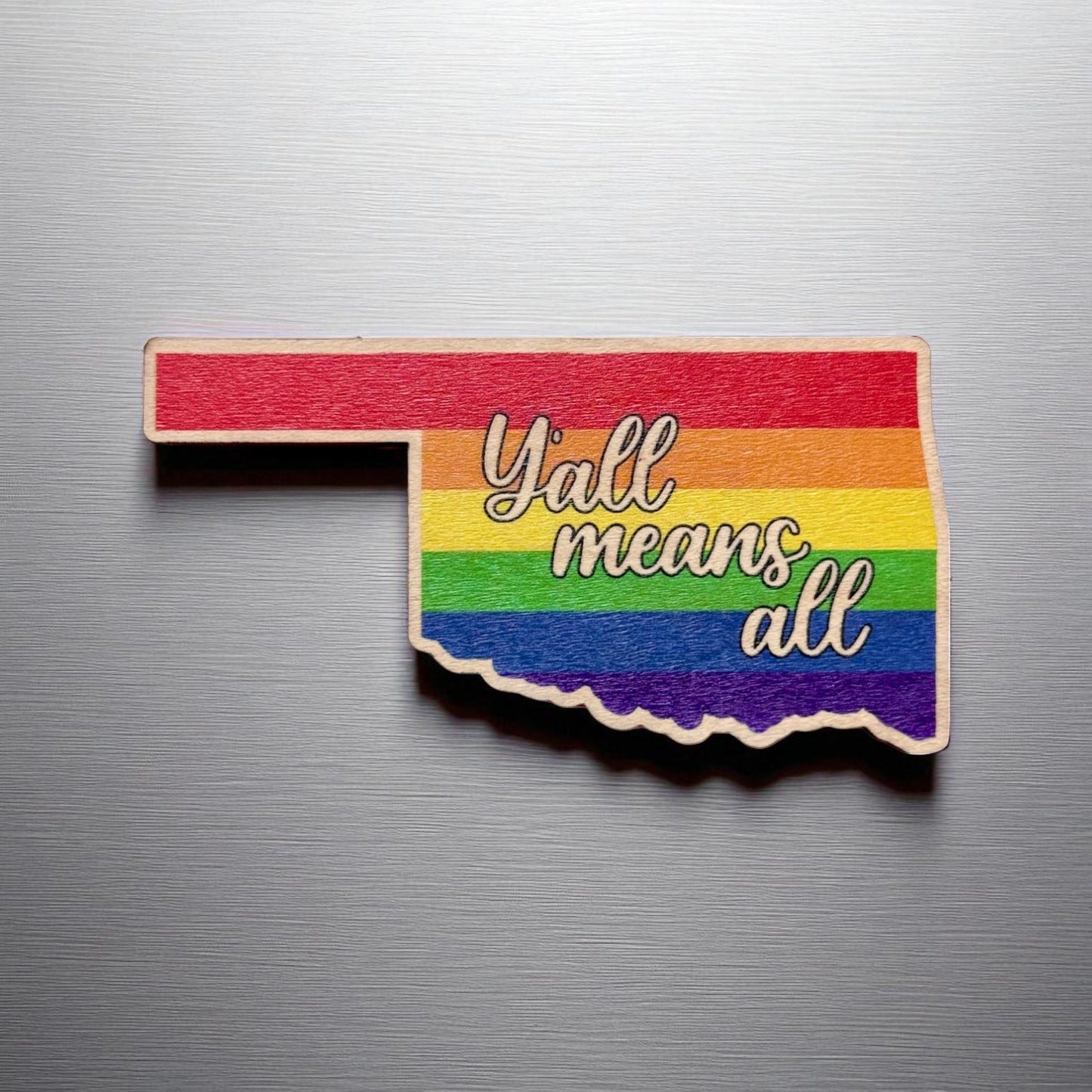 Straight-on view of Oklahoma-shaped rainbow wood magnet with bold message celebrating inclusivity and state pride