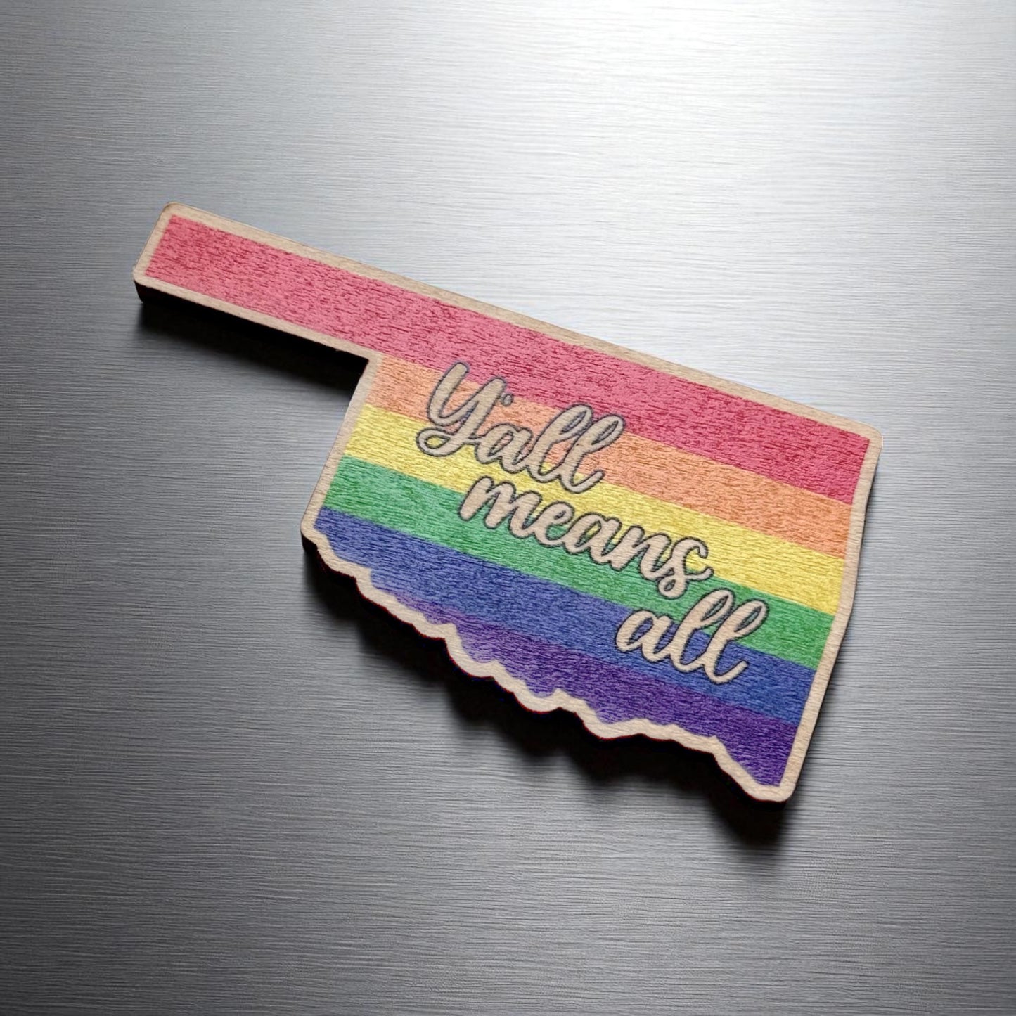 Angled view of Y’all Means All Oklahoma wood magnet on stainless steel fridge showing state cutout and colorful pride design
