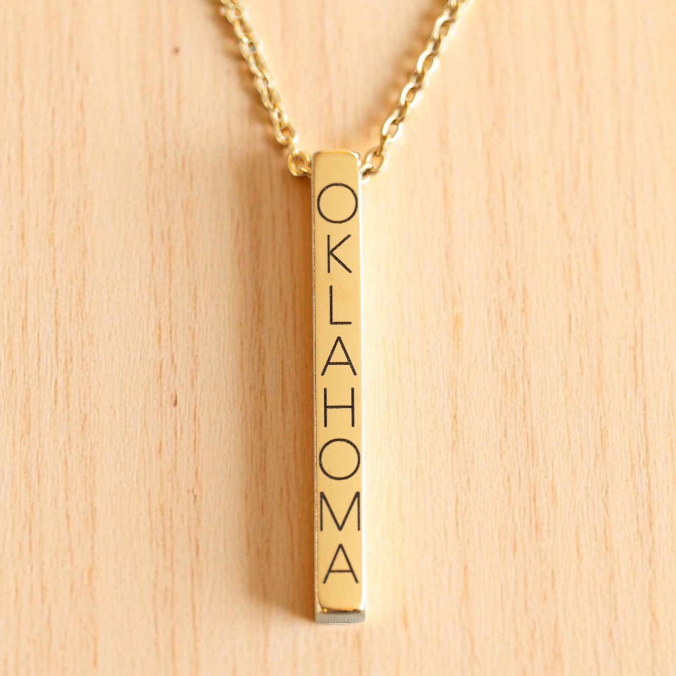 Minimal gold vertical bar necklace engraved with Oklahoma, hanging on gold chain.
