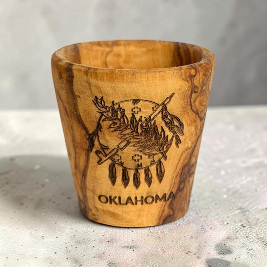Laser engraved Oklahoma flag olive wood shot glass standing on a tabletop, natural wooden Oklahoma souvenir and state gift with visible wood grain
