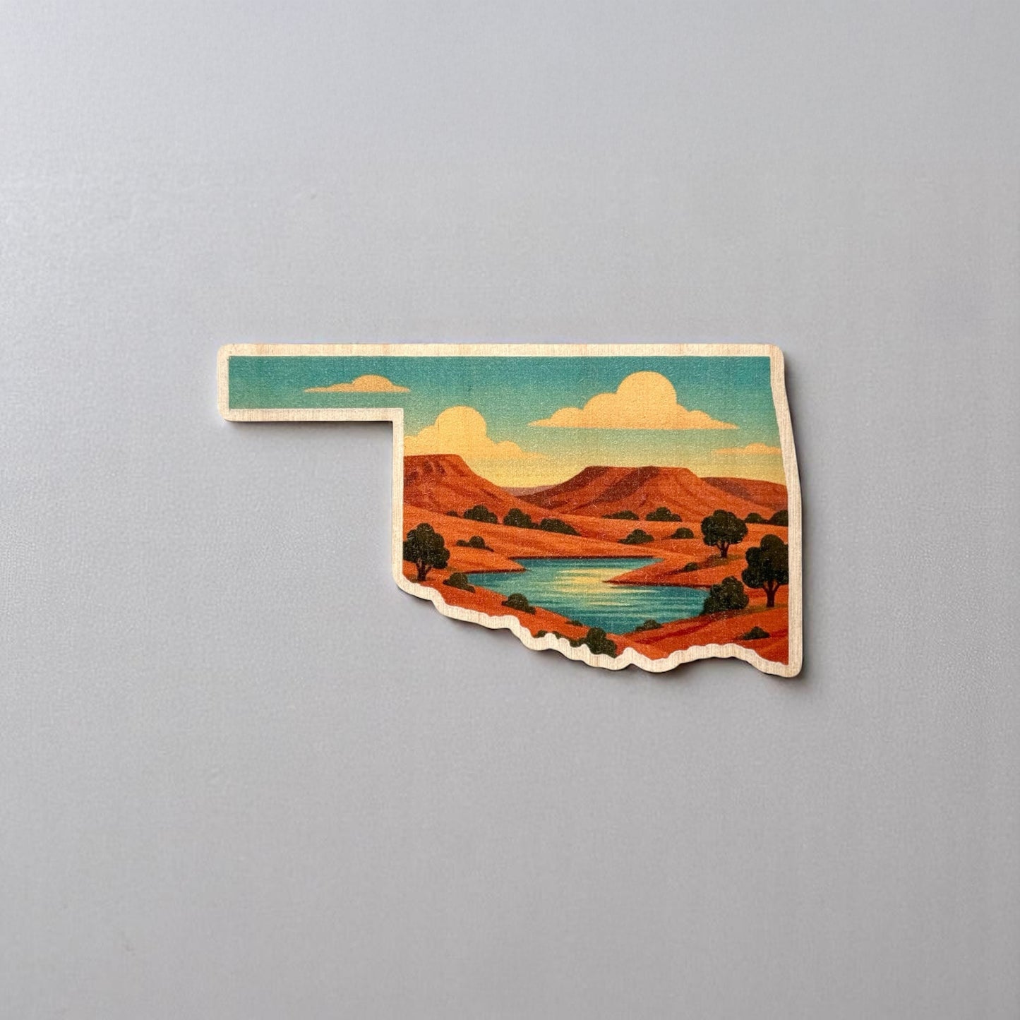 Illustrated Oklahoma wood sticker on gray background showing sunset-lit mesas, pine trees, and rivers in a desert landscape