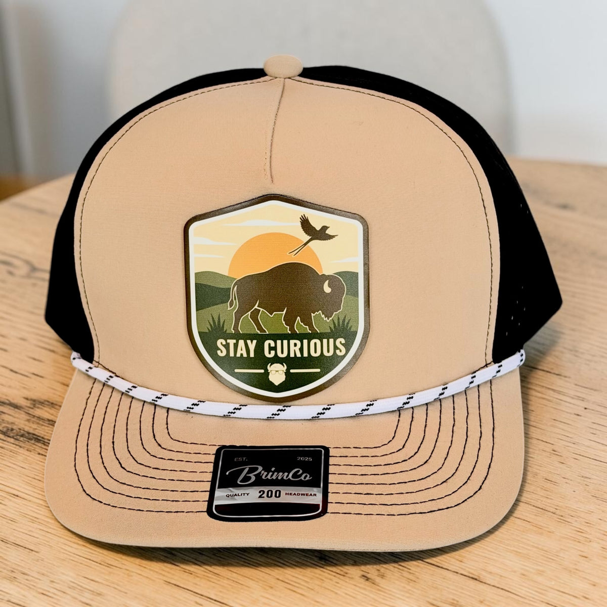 Close front view of tan and black performance golf hat with Stay Curious bison patch featuring sunset, Oklahoma scissor-tailed flycatcher, landscape artwork, and rope detail