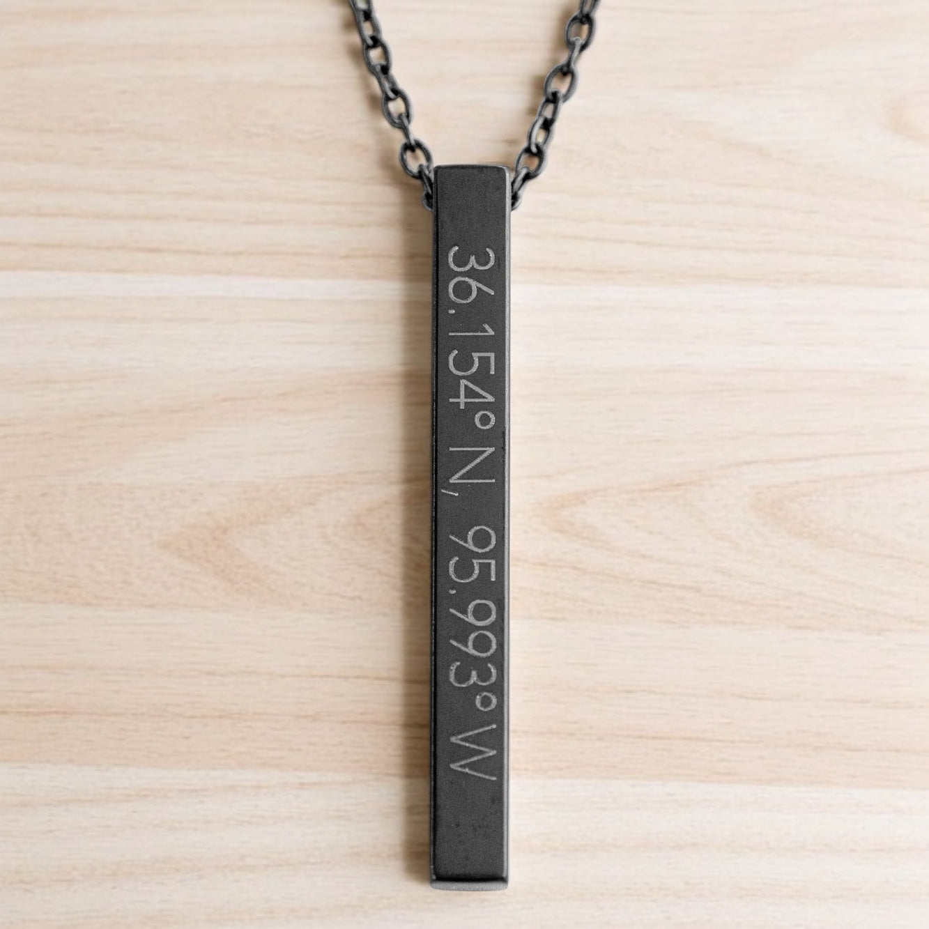 Matte black vertical bar necklace with Tulsa coordinates 36.154°N, 95.993°W, displayed on a light wood tabletop.
