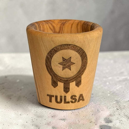 Laser engraved Tulsa flag olive wood shot glass standing on a tabletop, wooden Tulsa Oklahoma souvenir with TULSA lettering and visible natural wood grain
