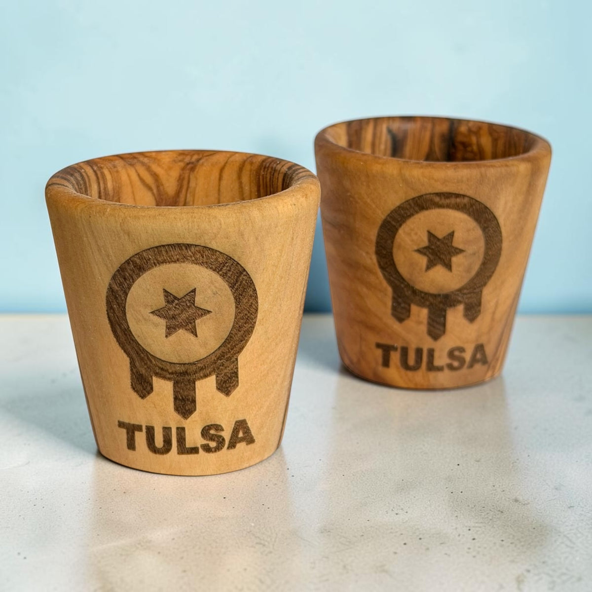 Pair of olive wood Tulsa shot glasses with laser engraved Tulsa flag designs, natural wooden Tulsa Oklahoma souvenirs showing variations in wood grain and color