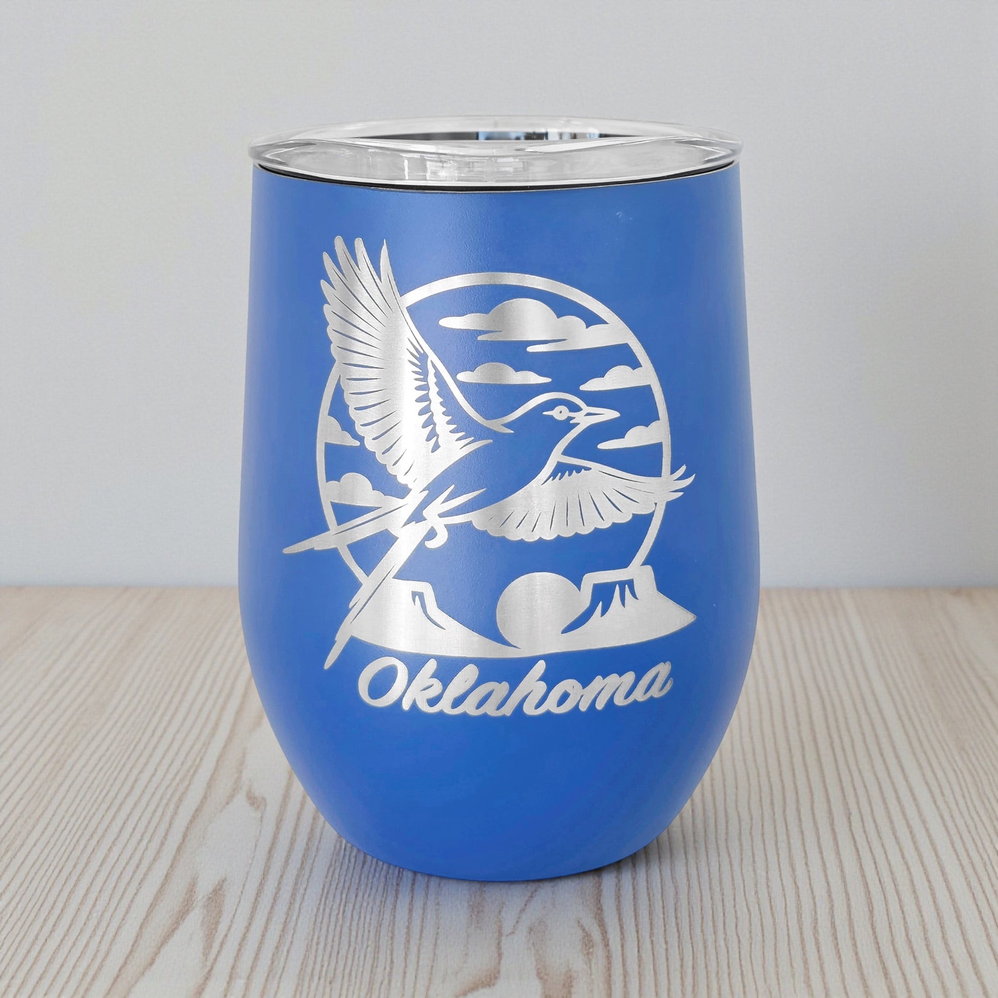 Blue 12oz wine tumbler with a laser etched Oklahoma scissortail flycatcher design, shown on a wood tabletop with a neutral background. This insulated stemless wine tumbler is a functional Oklahoma souvenir, ideal for wine and mixed drinks, and commonly purchased as a gift by Oklahoma locals and Route 66 travelers.