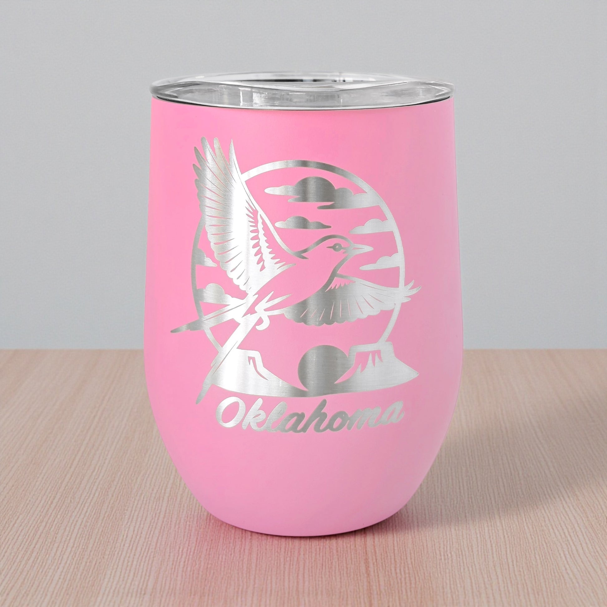 Pink 12oz wine tumbler featuring a laser etched Oklahoma scissortail flycatcher design with clouds and rolling landscape details, photographed on a light wood surface. This insulated stainless steel wine tumbler includes a clear lid and is a popular Oklahoma gift, state pride drinkware item, and Route 66 souvenir for wine, cocktails, and everyday use.
