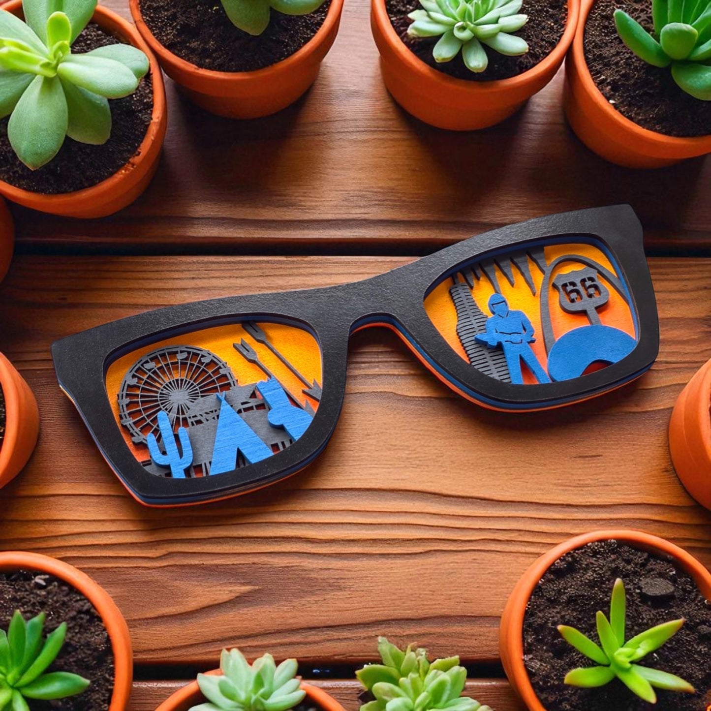 Black wooden Route 66 wall art sunglasses placed among terracotta potted succulents, showcasing laser-cut travel icons from across Route 66 with a vibrant orange backdrop.