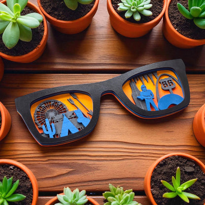 Black wooden Route 66 wall art sunglasses placed among terracotta potted succulents, showcasing laser-cut travel icons from across Route 66 with a vibrant orange backdrop.