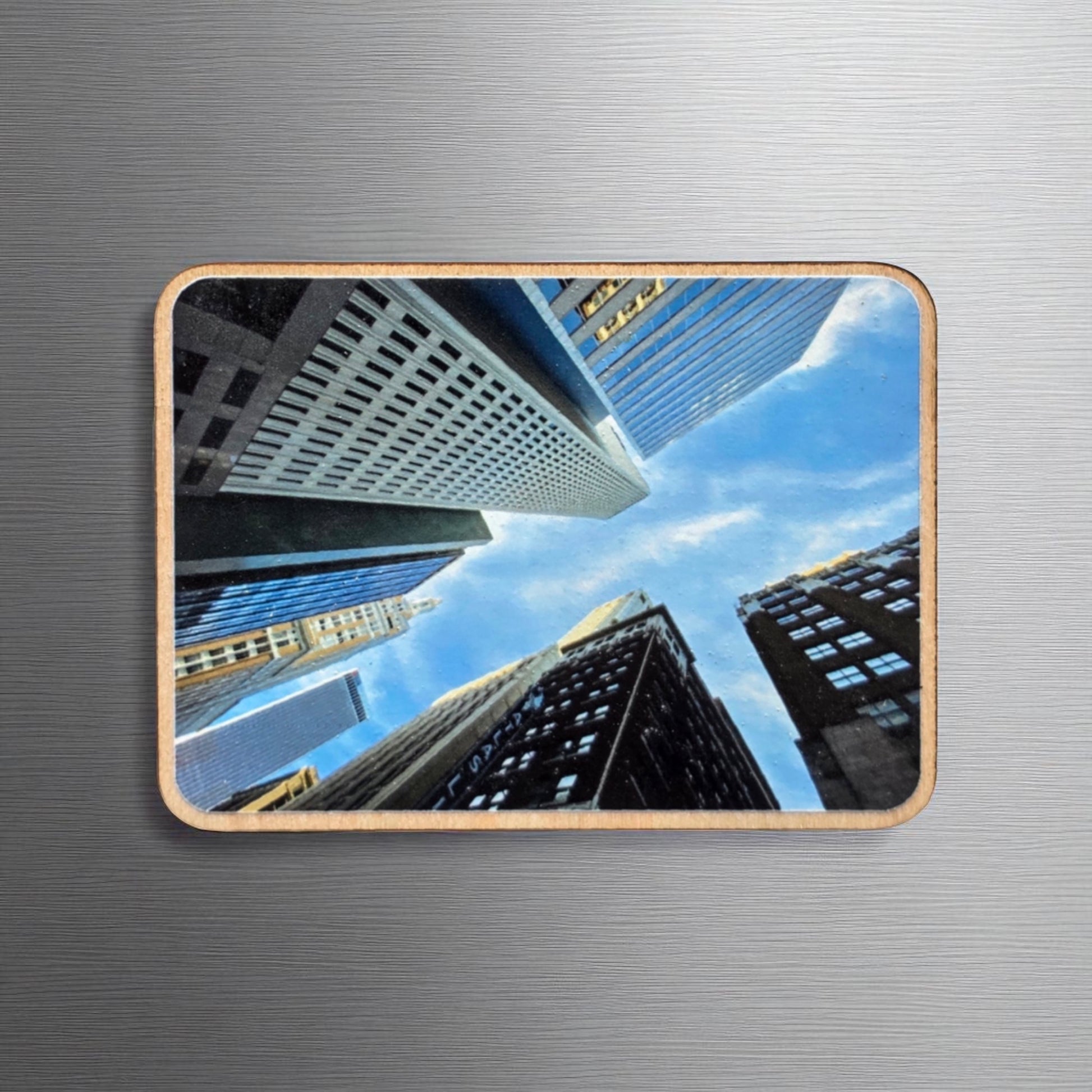 Wooden photo magnet featuring a dramatic upward view of Tulsa, Oklahoma skyscrapers against a blue sky, displayed on a metallic surface. The perspective captures the height and variety of the city’s downtown architecture.