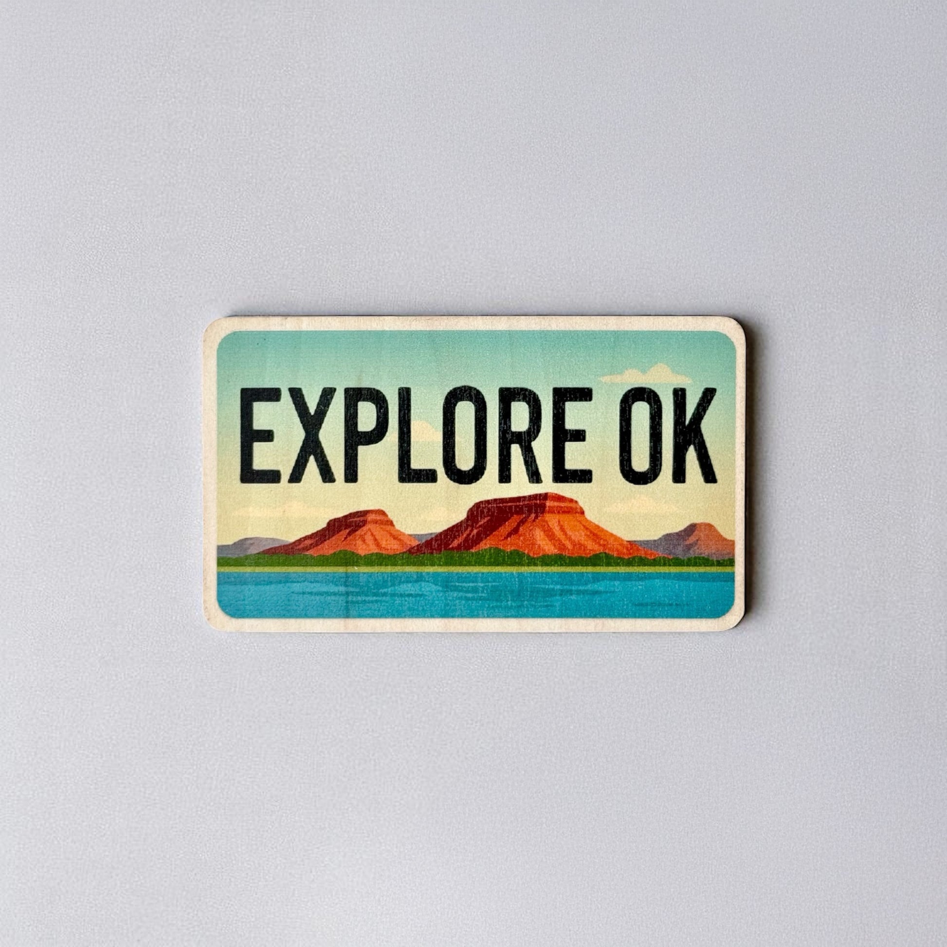 Vintage-style wood sticker that says “Explore OK” with red mesas and bright blue water, inspired by Oklahoma’s natural beauty.