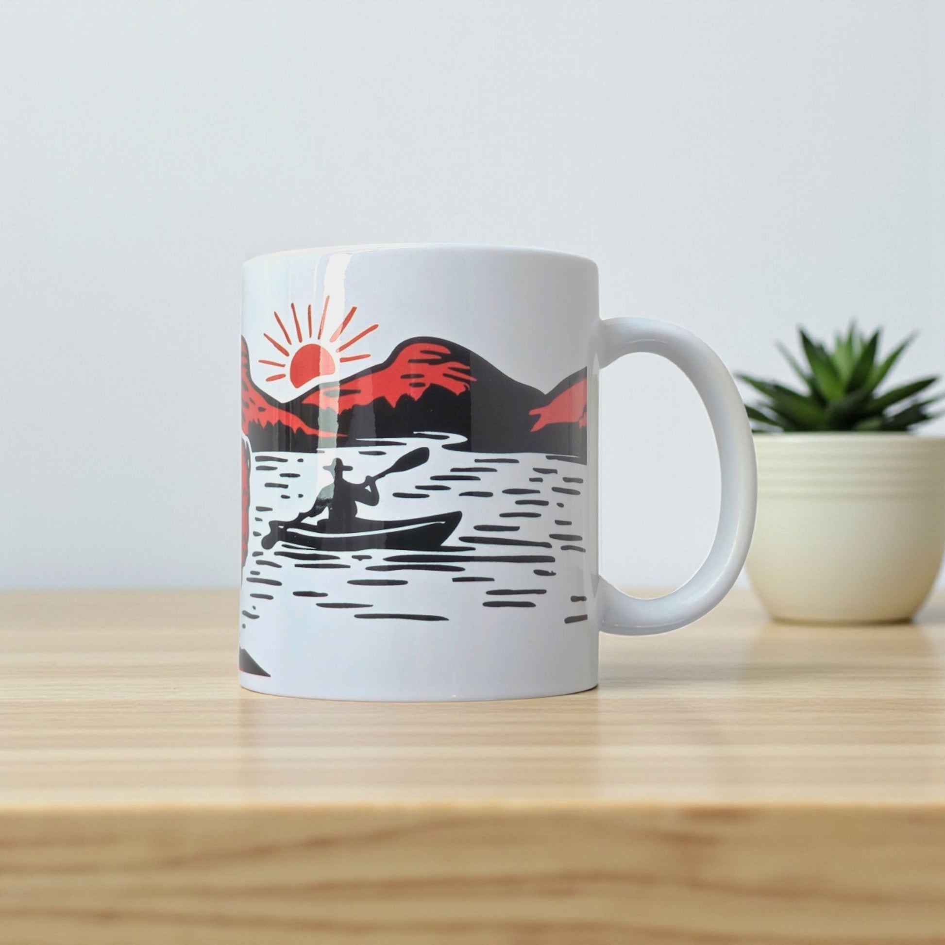 Adventure-themed Oklahoma mug featuring kayaker and mountain sunrise, displayed on wooden table.
