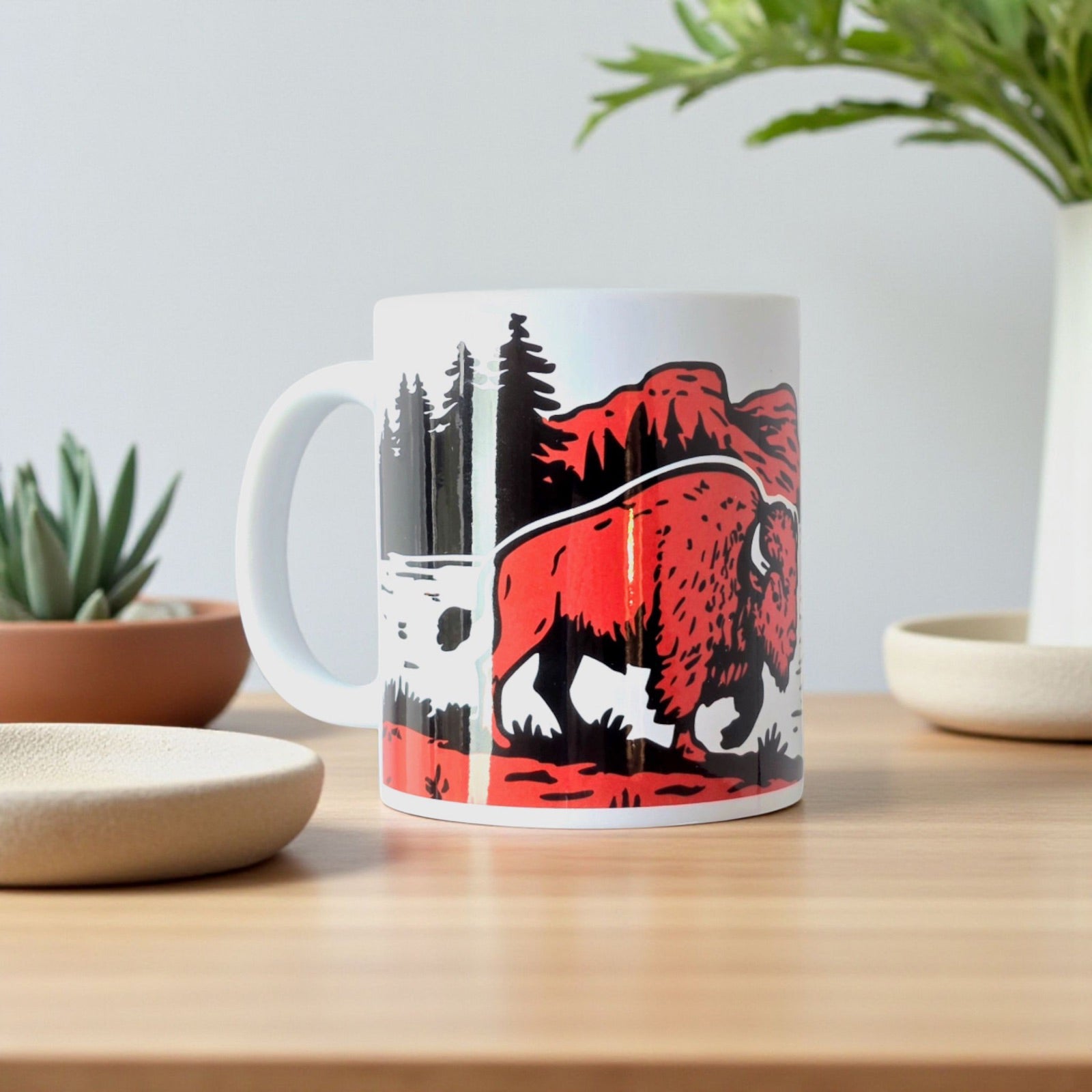 Oklahoma bison adventure mug with forest and mountains design, styled with plants on table.