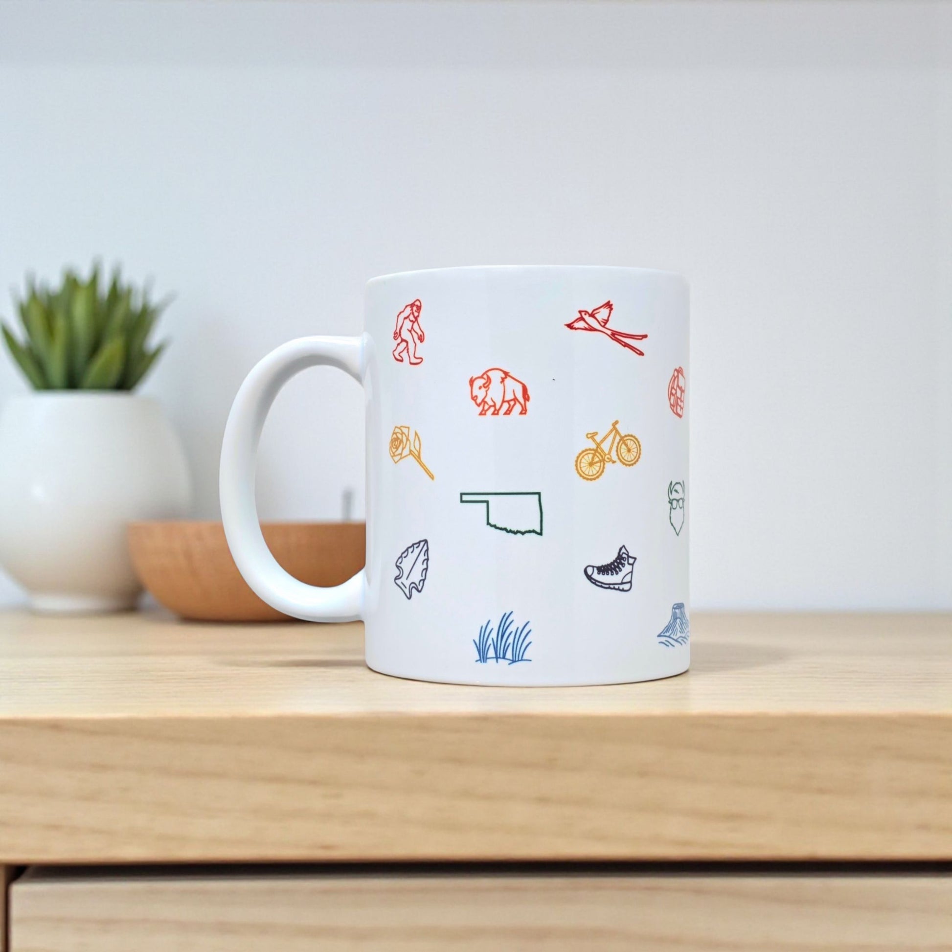 Oklahoma icons coffee cup featuring colorful state symbols, displayed with plate on wooden surface.