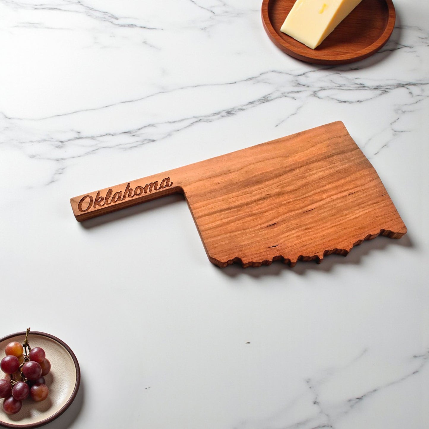 Oklahoma cherry wood serving board on marble surface with grapes and cheese nearby.