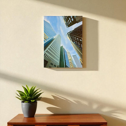Vertical 12x16 inch Tulsa skyscraper canvas wall art shown hanging on a light-colored wall above a wooden table with a small green plant, bathed in afternoon light