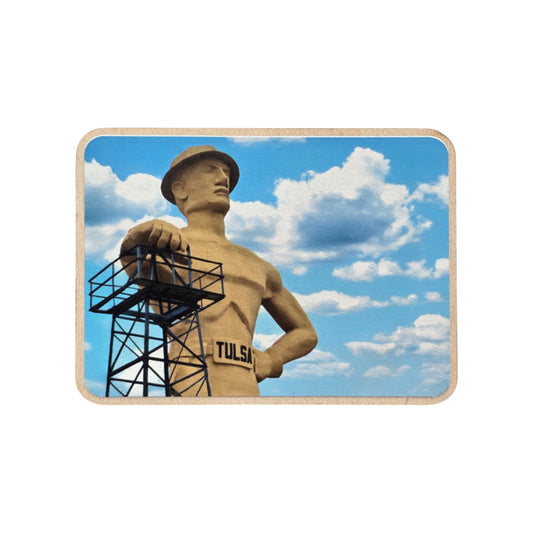 Wood photo magnet featuring the Tulsa Golden Driller statue against a blue sky with fluffy clouds. The statue stands tall with one hand resting on a black oil derrick, wearing a belt labeled “TULSA.” Displayed on a white background.