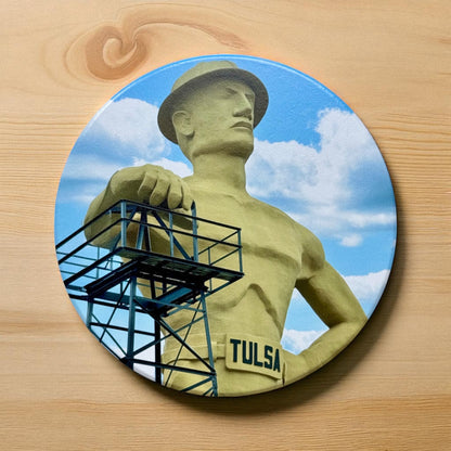 Colorful round ceramic coaster featuring Tulsa’s Golden Driller statue under a blue sky with white clouds, placed on a light wood surface