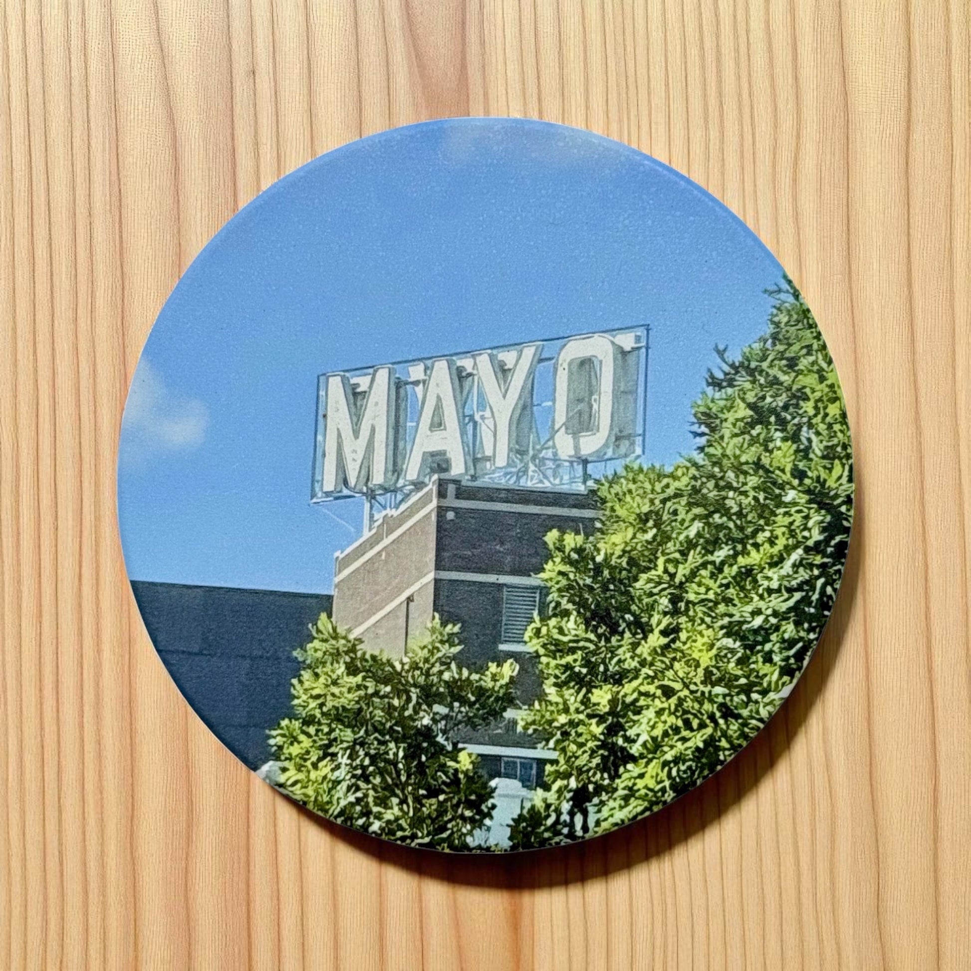 Ceramic drink coaster with a vivid photo of the Mayo Hotel rooftop sign in Tulsa, Oklahoma, surrounded by green trees and a clear blue sky, placed on a wood grain surface
