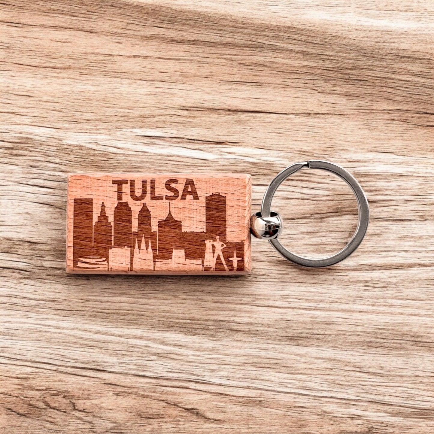 Tulsa skyline wood keychain displayed on a woodgrain background, featuring etched downtown buildings and the word “TULSA.”