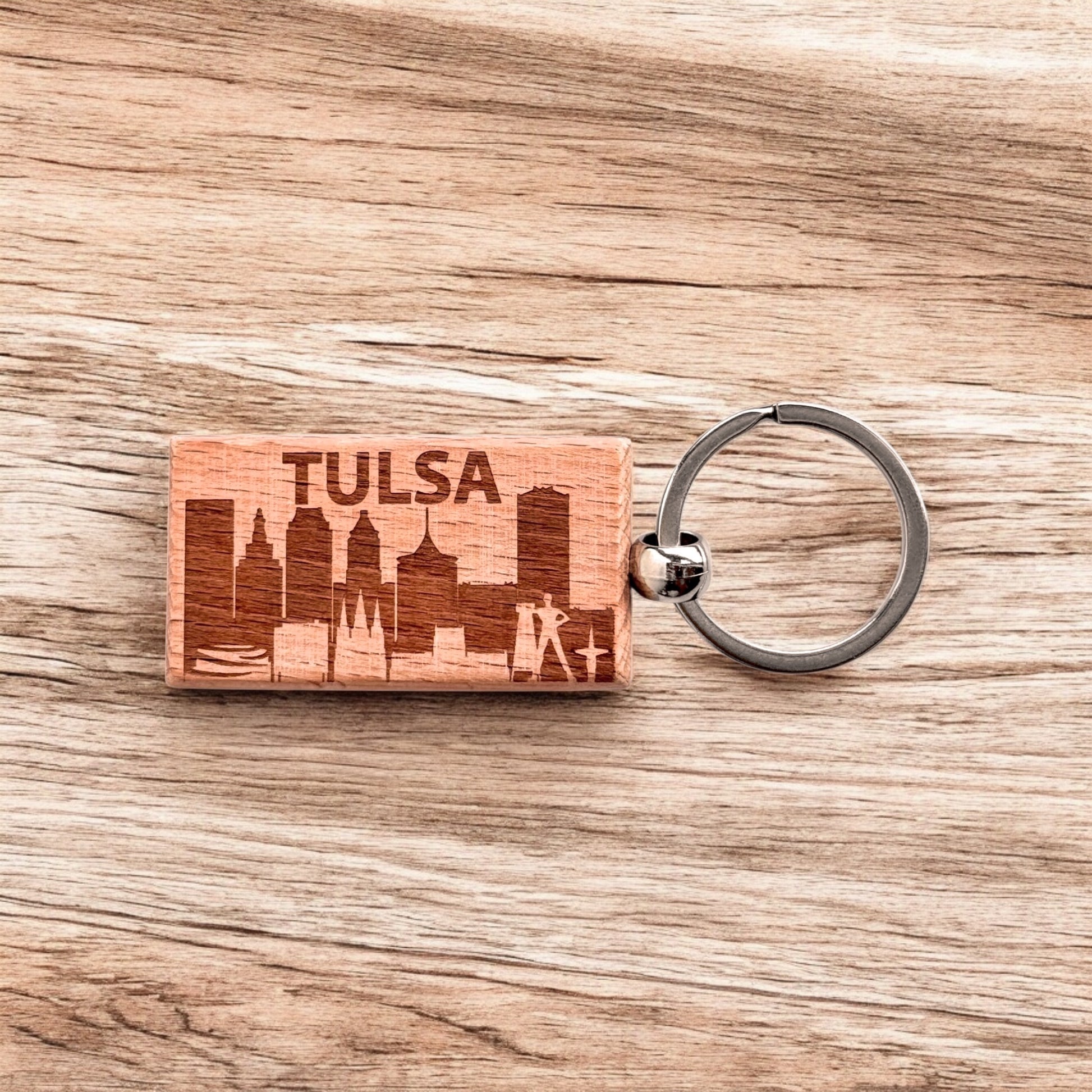 Tulsa skyline wood keychain displayed on a woodgrain background, featuring etched downtown buildings and the word “TULSA.”