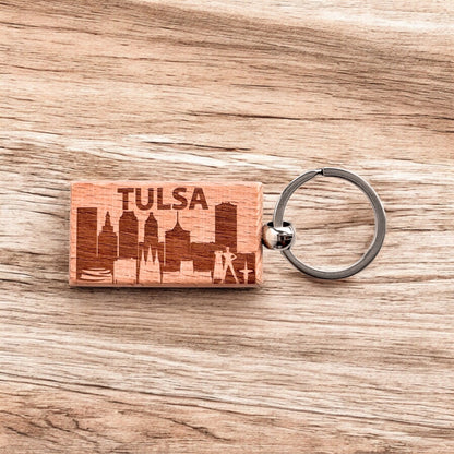 Tulsa skyline wood keychain displayed on a woodgrain background, featuring etched downtown buildings and the word “TULSA.”