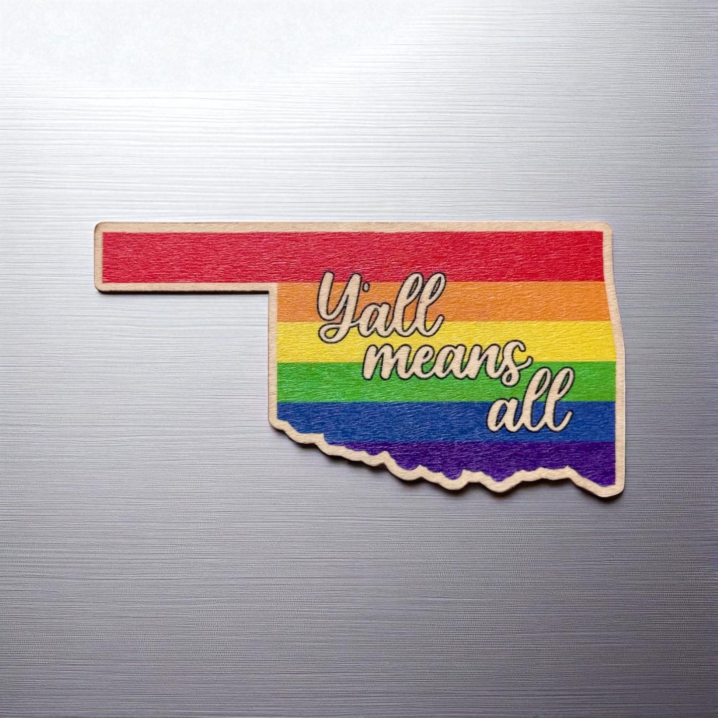 Flat view of rainbow-colored Oklahoma-shaped wood sticker reading “Y’all Means All,” applied to a metal surface