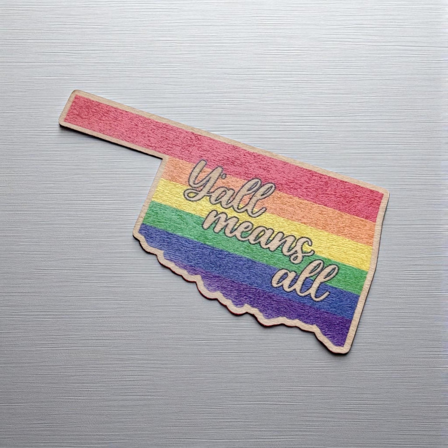 Wood sticker in the shape of Oklahoma printed with rainbow stripes and “Y’all Means All” message, placed on a brushed metal background