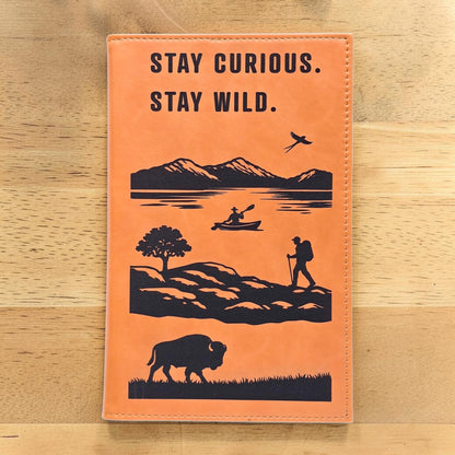 Adventure journal with Stay Curious Stay Wild quote on tan leatherette cover featuring mountain lake kayak hiker bird and bison artwork, travel notebook gift for hikers campers and road trip lovers in the USA