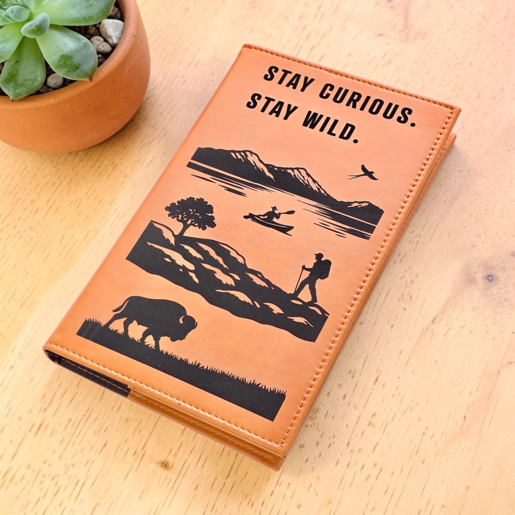 Angled view of tan adventure journal with Stay Curious Stay Wild cover design beside plant, leatherette travel notebook for journaling trips hiking memories and daily notes