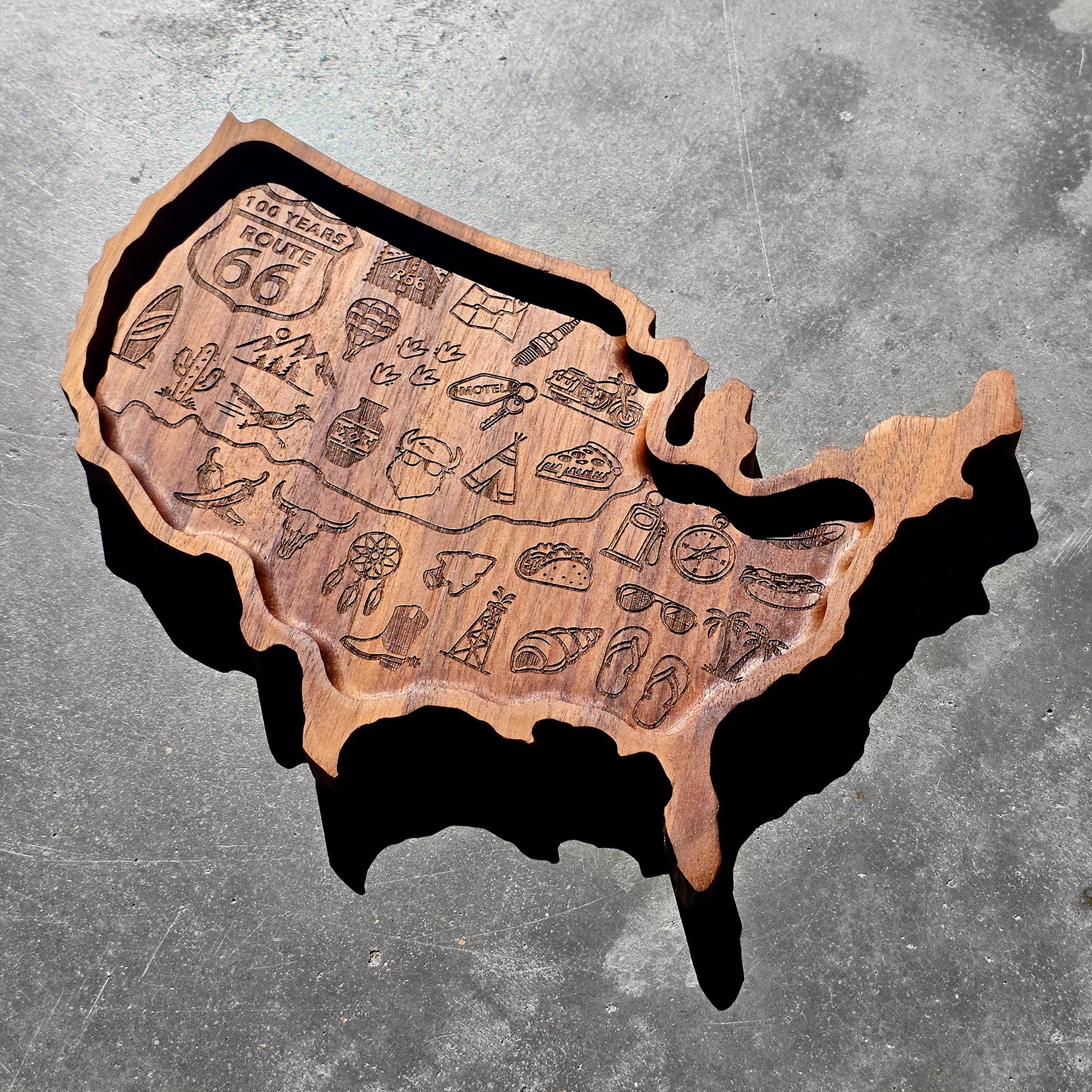Angled view of USA shaped wooden valet tray with engraved travel themed icons representing classic American road trips, western culture, Route 66 travel, and vintage roadside attractions