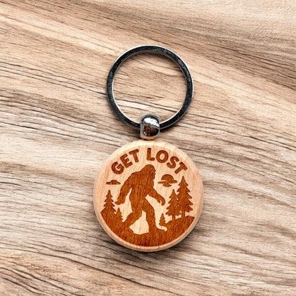 Laser engraved Bigfoot get lost wooden keychain resting on wood surface, outdoor themed wood keychain with forest design for hiking camping gift or nature souvenir