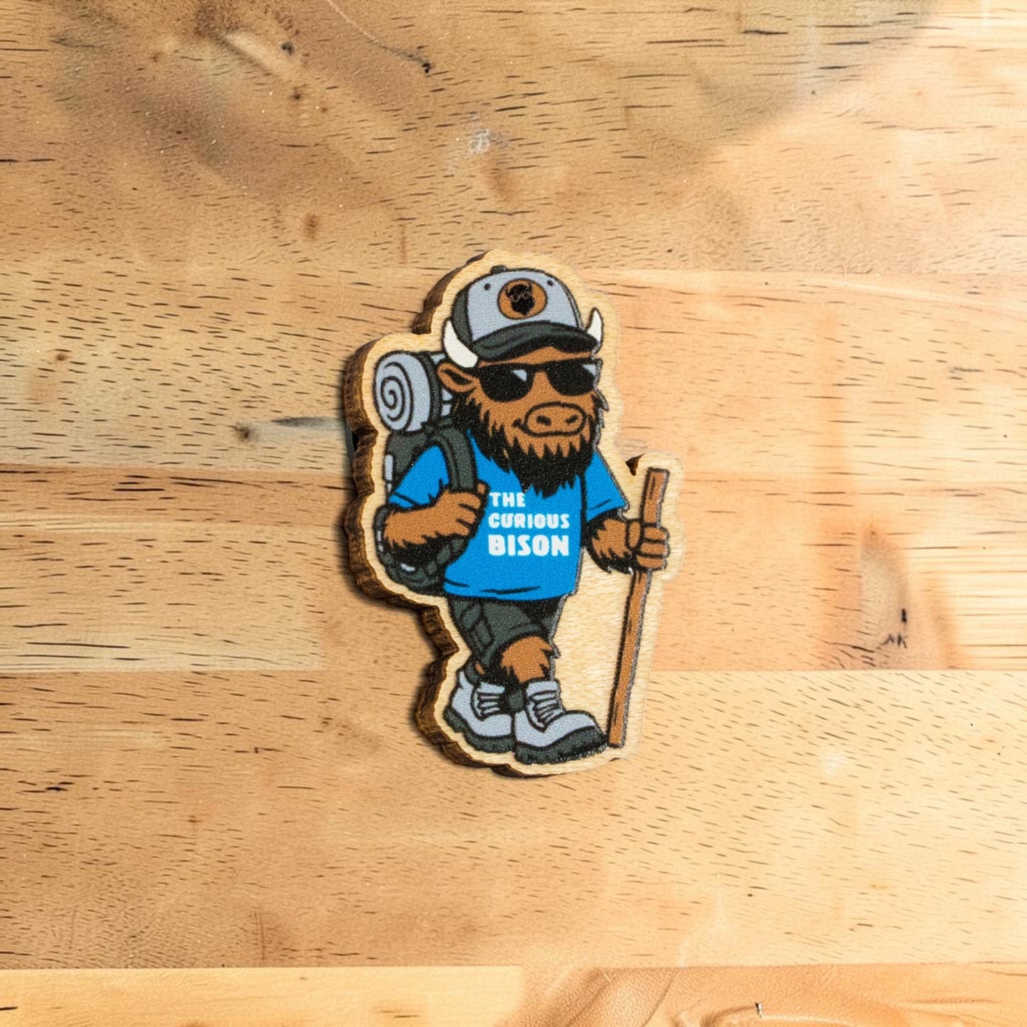 Wooden bison lapel pin made from Eco Wood Birch featuring hiking mascot with backpack, sunglasses, walking stick, and blue shirt on wood table