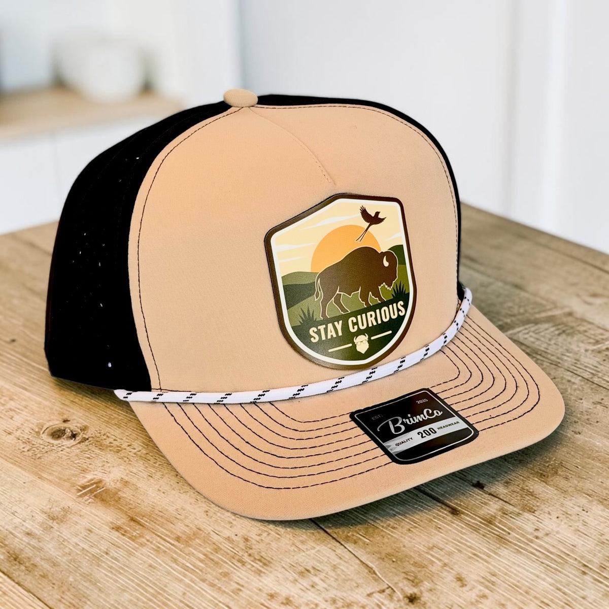 Angled view of tan and black bison performance golf hat showing Stay Curious outdoor patch, rope accent, precurved bill, and laser-perforated side panels