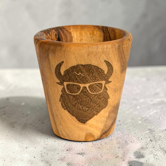 Laser engraved bison olive wood shot glass standing on a tabletop, wooden buffalo souvenir featuring a bison wearing glasses and natural wood grain