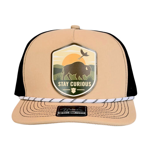 Front view of tan and black bison performance golf hat with full-color Stay Curious patch featuring a bison, scissor-tailed flycatcher, sunset landscape, and rope cap detail